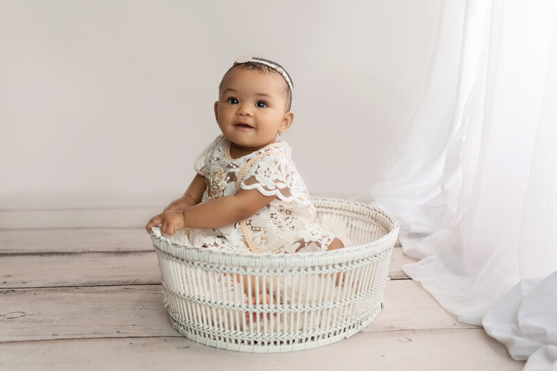 A toddler girl in a white lace dress sits in a wicker basket in a studio under a window of a Lafayette Baby Milestone Photographer studio