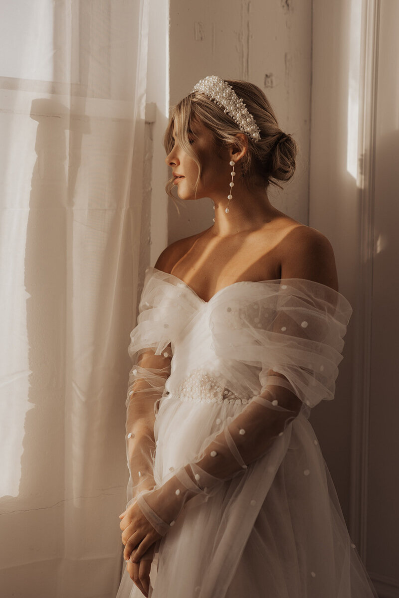 Bride by window in soft light, wedding shoot.