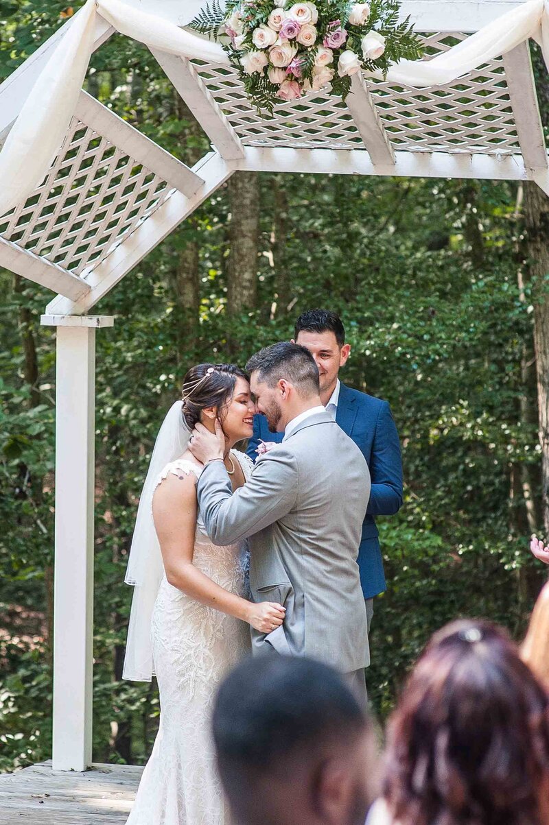 Bride in lace and beaded wedding gown with a plunging deep v neck wedding dress embraces a groom with light grey suit under a lattace arbor
