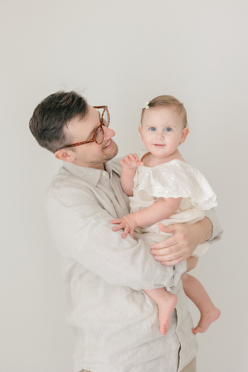 A dad holding his one year old daughter looking at her lovingly in all white studio in NJ