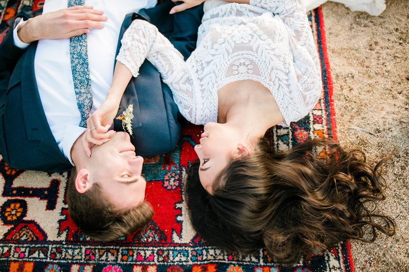 the couple lays on a red patterned boho rug. the bride touches the grooms face