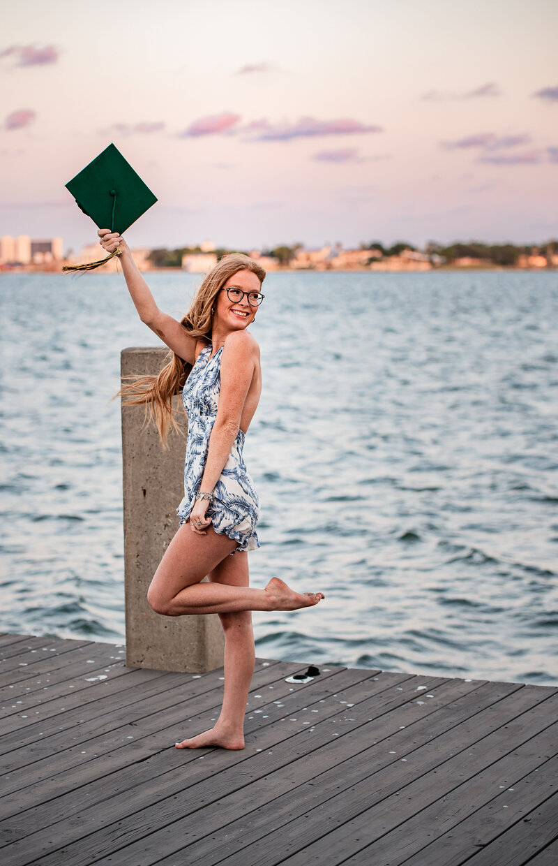 A senior stands on a boat dock in Galveston and holds her cap in the sky in celebration.