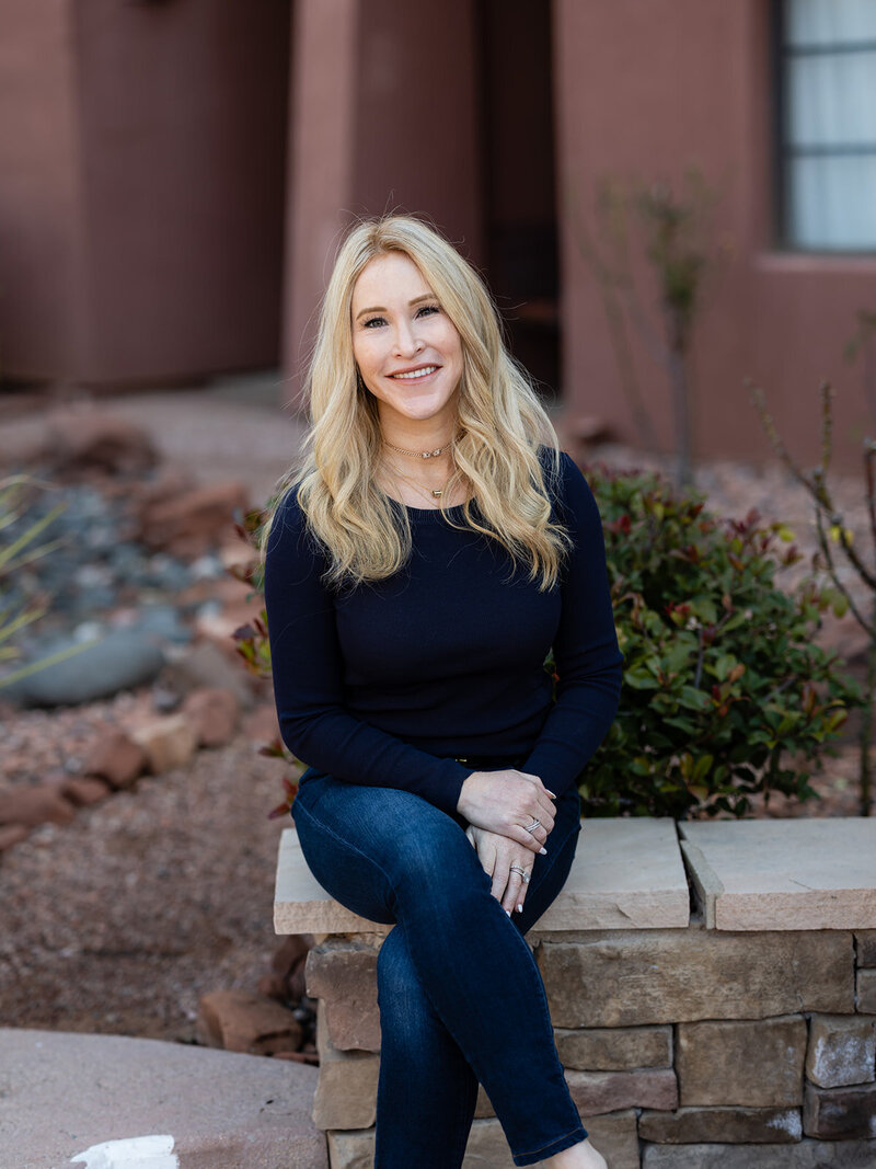 this is Lindsay Lovell, a real estate investor and coach sitting on a concrete bench and smiling at the camera, wearing a blue long sleeves and jeans