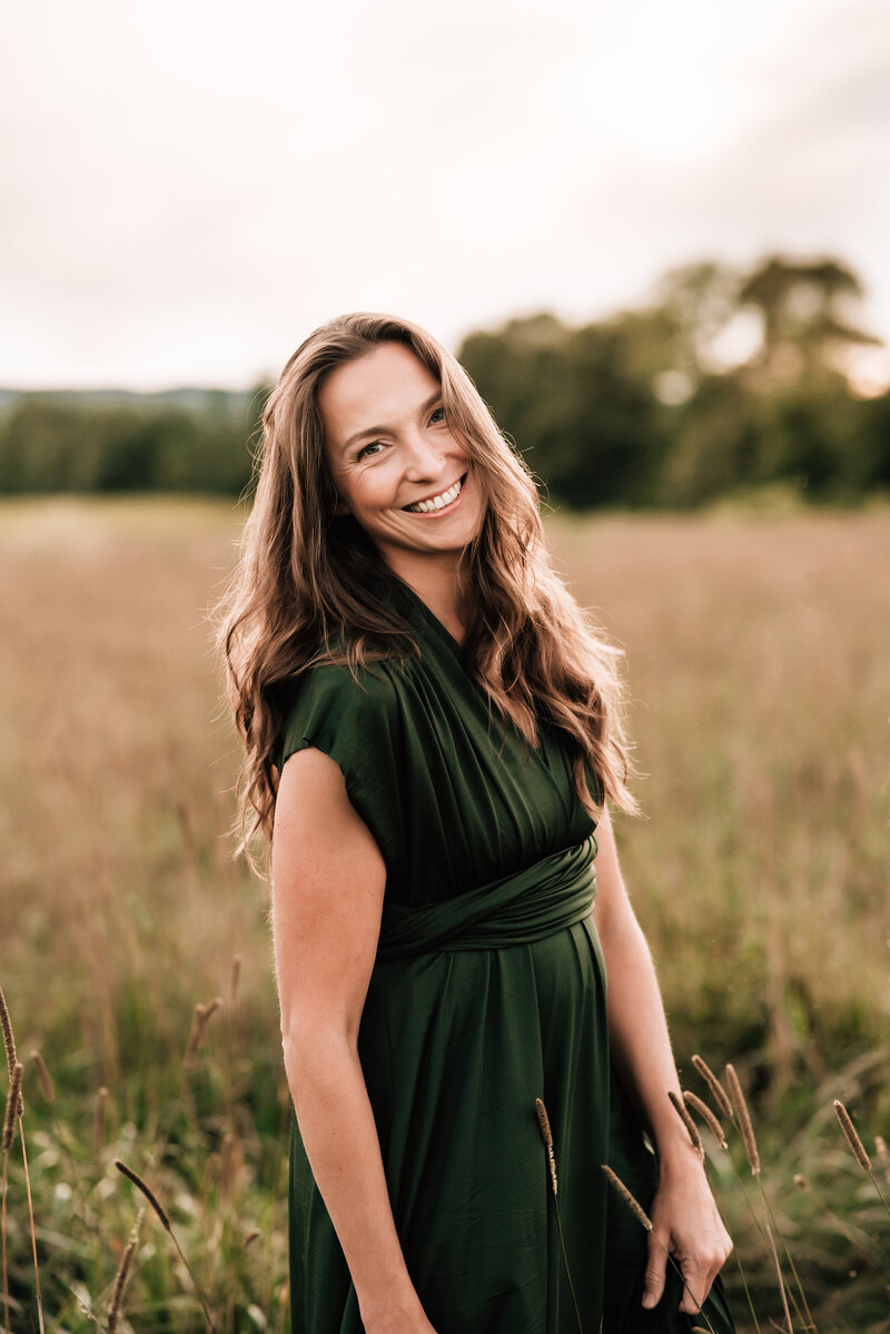 Katie Lintern in a tall grass field during sunset wearing a long green dress looking at the camera and smiling