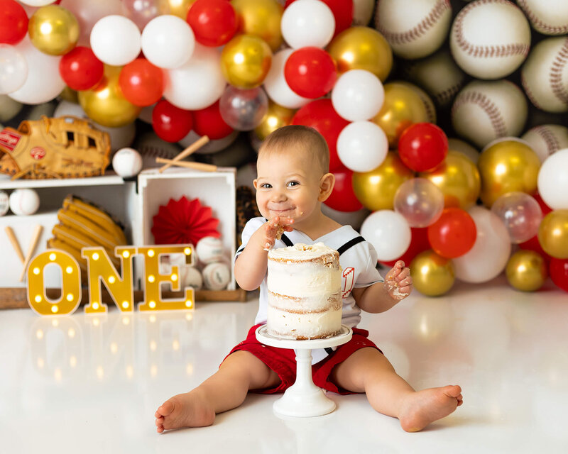 A little boy sits behind a cake smiling, in front of a baseball background