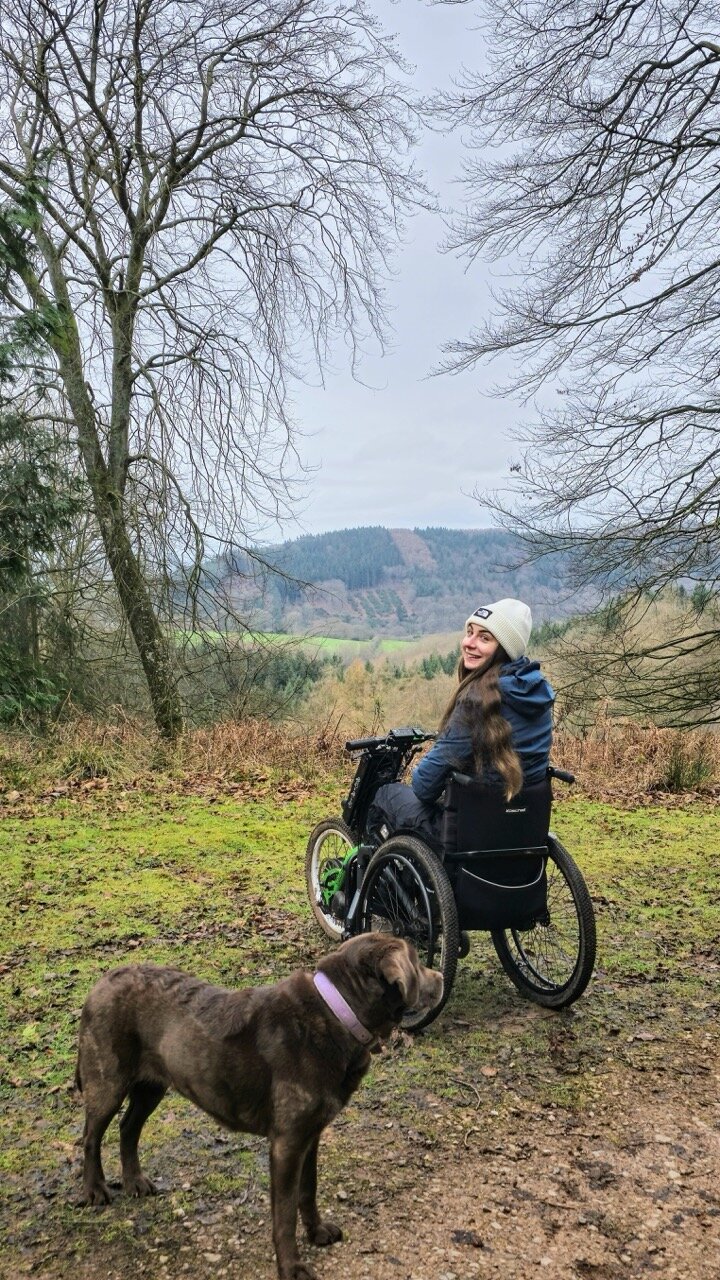 Bethany Handley in a wheelchair on a forest path with her dog, overlooking a scenic wooded valley.