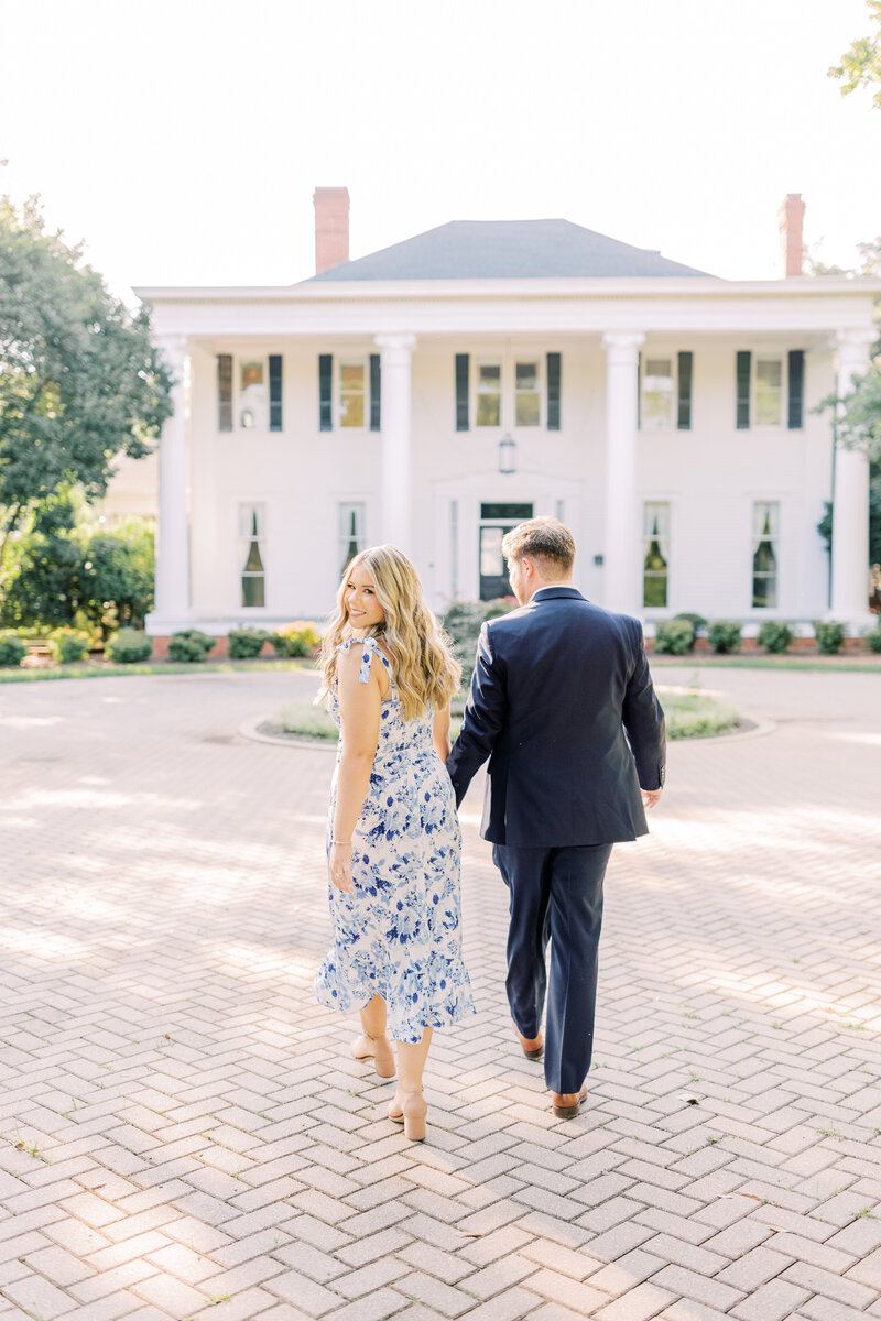 Woman holding hands with man looking back at camera in front of a white mansion