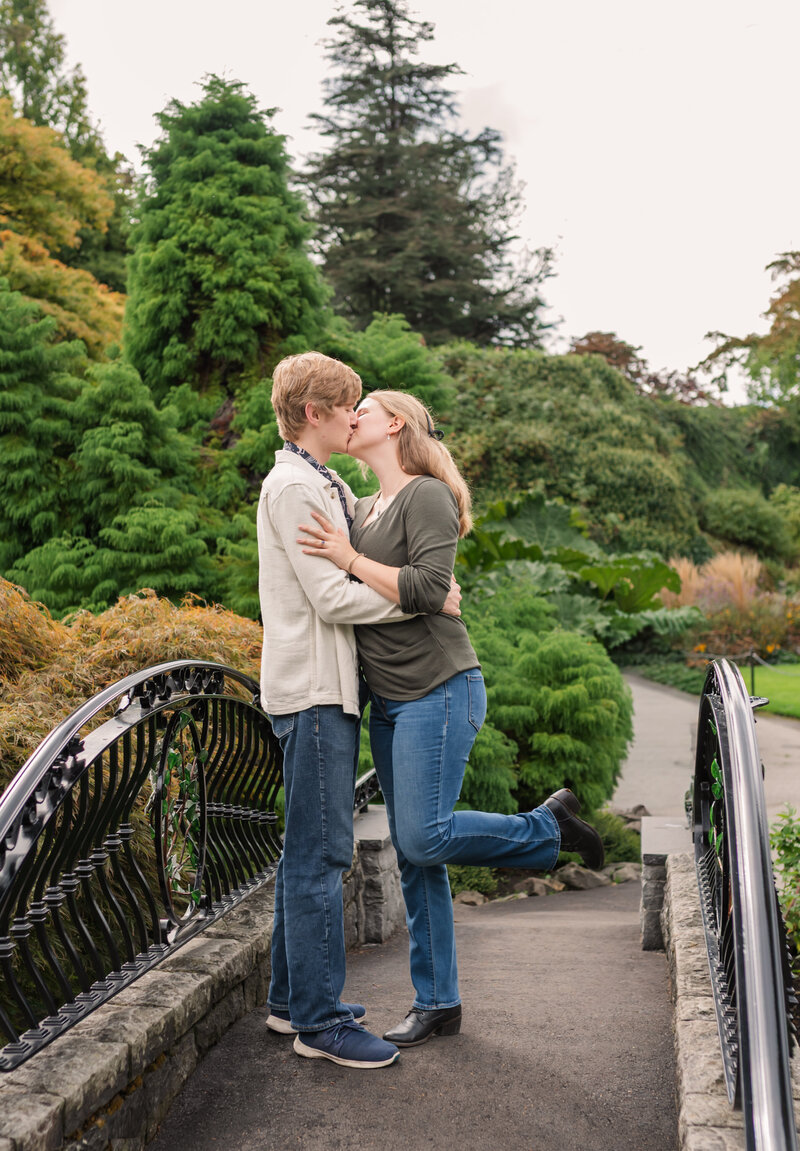 newly engaged couple kissing on a bridge at Queen Elizabeth Park. Girls left foot extended out while couple kiss.