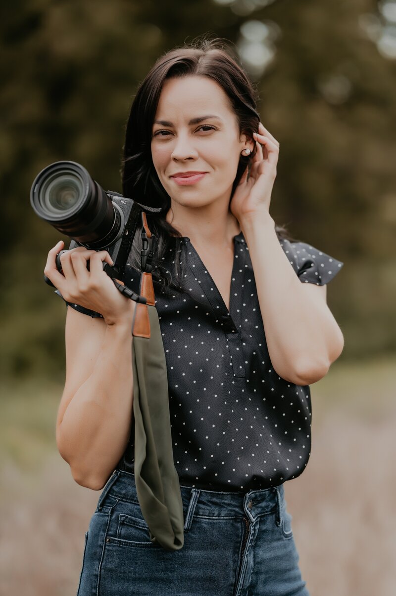Best wedding photographer and videographer in London, ON Ashlee Ellison is smiling and holding her camera as she poses for a headshot. She is wearing a black blouse with tiny white polkadots and pushing her hair behind her ear. She is in a field with evergreens in the distance.