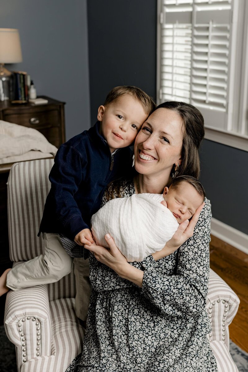 mother holds newborn wrapped and white with her toddler son standing beside her holding on to her