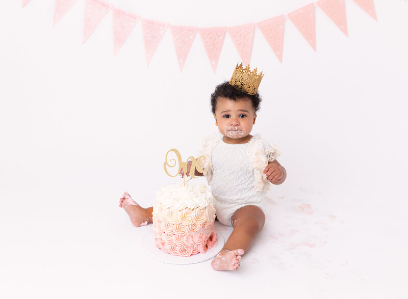 Rochel Konik Photography | Top Brooklyn Cake Smash Photographer captures baby girl in white lace romper and floral crown getting messy eating her first birthday cake in a studio cake smash session. Baby has icing on her hands , feet, and legs and is looking up at the camera.