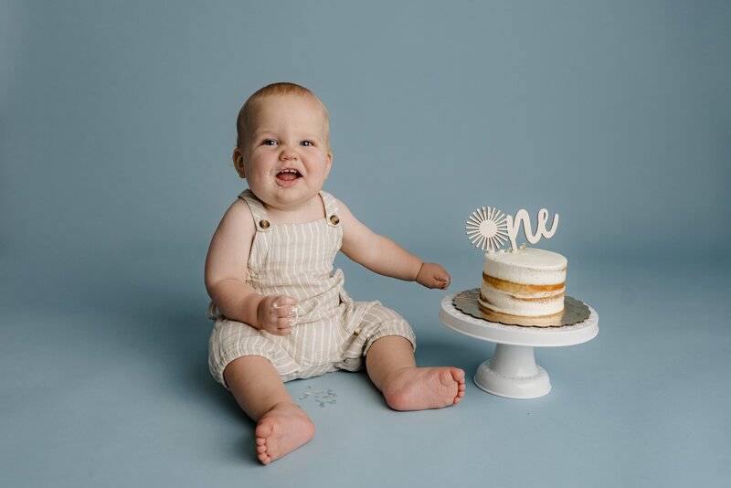 simple cake smash photo session of a little boy dressed in a neutral onsie with his cake on a stand beside him and sitting on a blue background