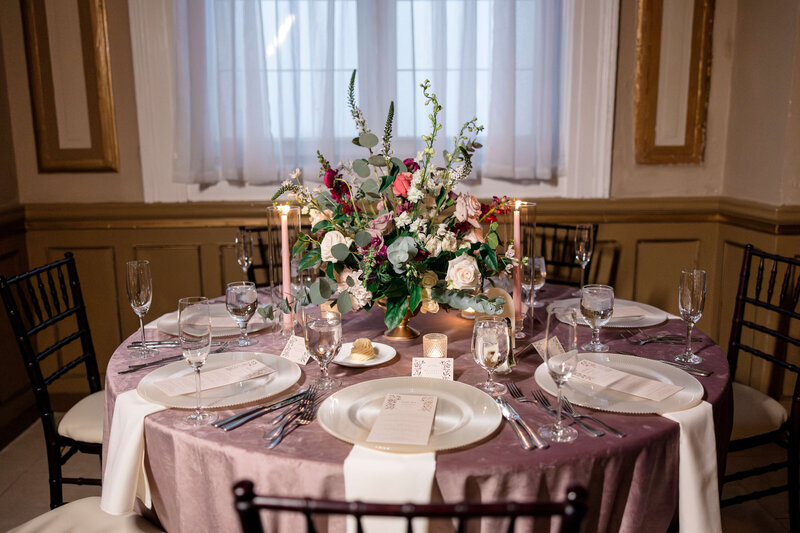 An elegant table setting features a large floral centerpiece with pink and white flowers and greenery. The table is set with plates, menus, wine glasses, and silverware. Tall candles are placed on either side of the centerpiece. Soft lighting prevails.