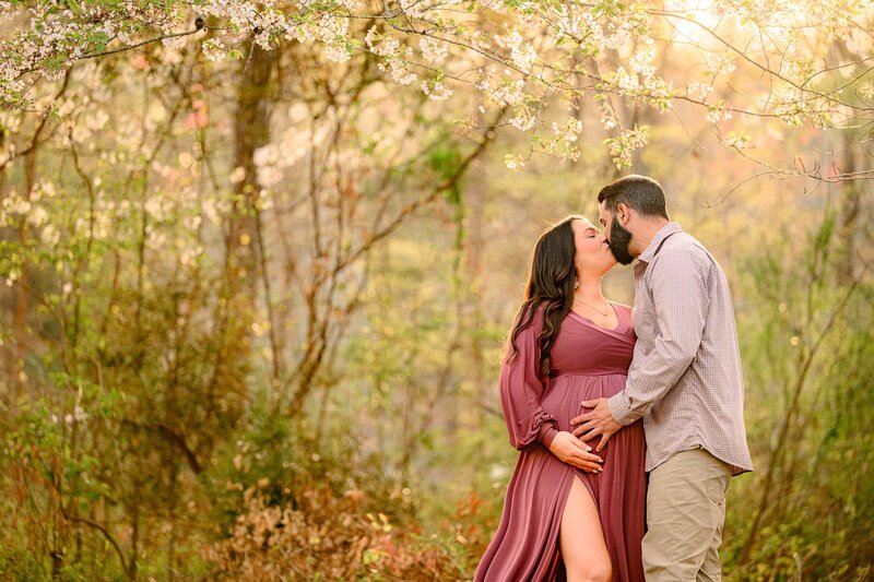Charlotte Area couple kisses and celebration of their upcoming arrival of their new baby girl during the maternity photo session where Mom is wearing a rose pink dress and dad is wearing a button-down shirt and khakis