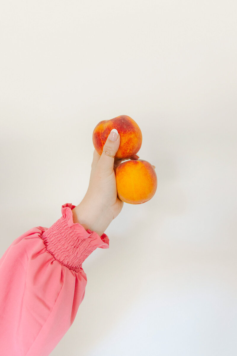 Mollie Mason's arm with a pink sleeve holding up 2 peaches in front of a white wall