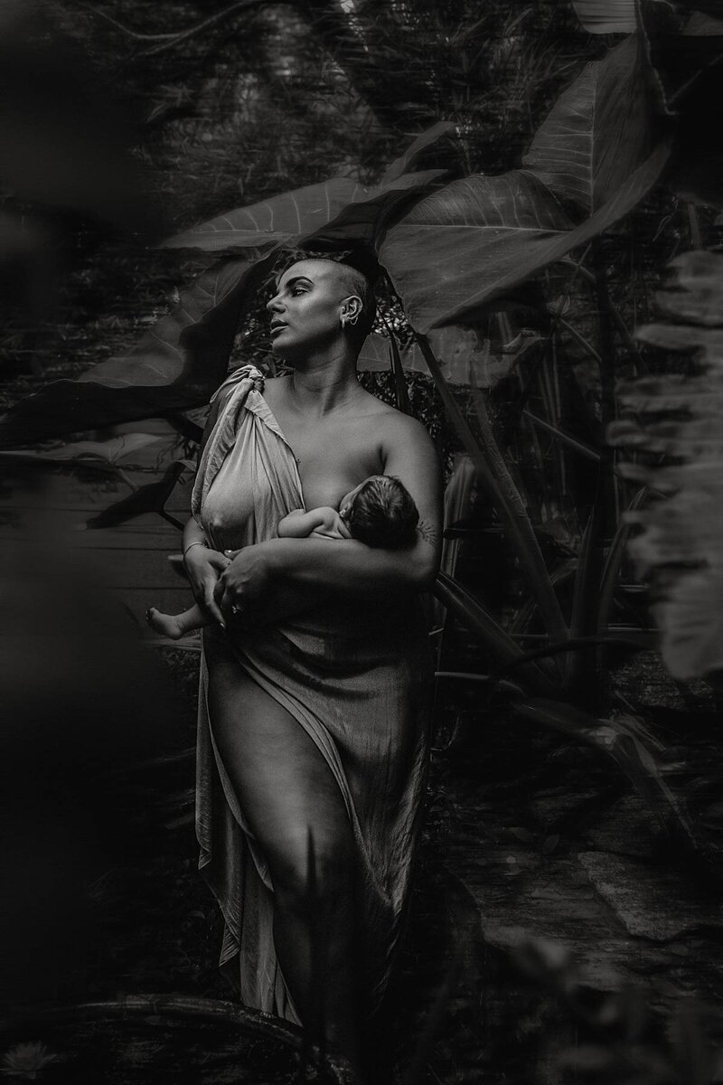 Emotive outdoor motherhood photography showing a mother nursing her baby among tropical plants.
