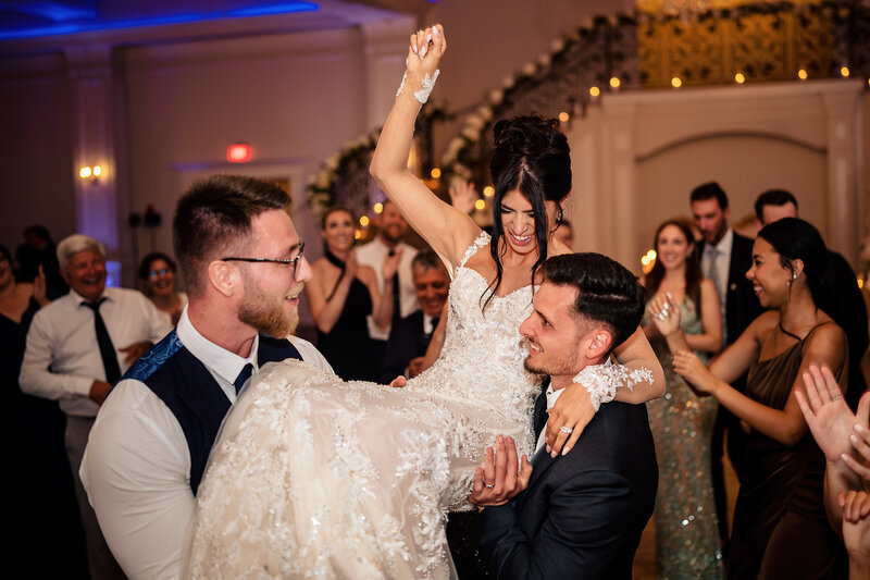 Bride exuberantly raised in a chair by groom and guest, her joy infectious amid a cheering crowd at a beautifully lit reception hall, encapsulating a moment of pure wedding bliss