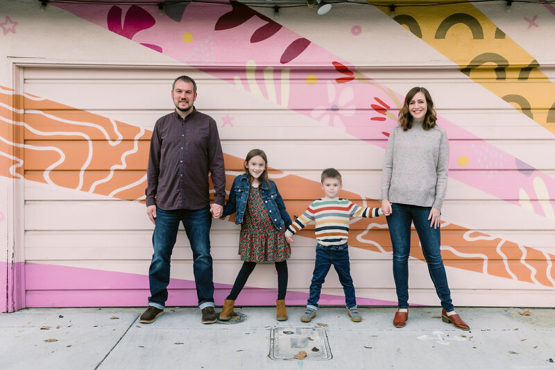 Parents and kids stand side by side holding hands in front of a colorful mural
