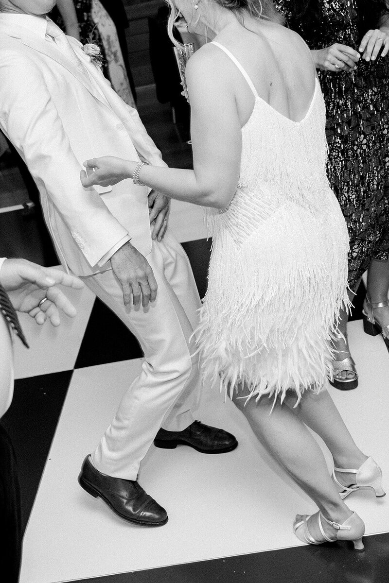 Black and white film-style moment of bride in feather mini dress dancing with groom on checkered floor at a San Francisco wedding