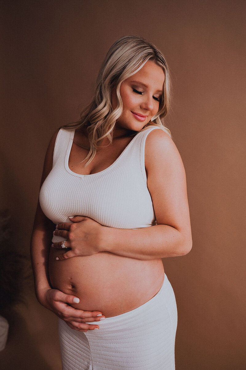 Pregnant woman wearing bralette matching set holding belly and smiling with eyes closed in a studio