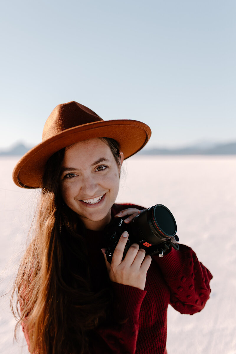 Orlando Wedding Photographer Jo from Four Loves Photo and Film looks into the camera while smiling and wearing a wide brimmed hat while salt flats are blurred in the background behind her.