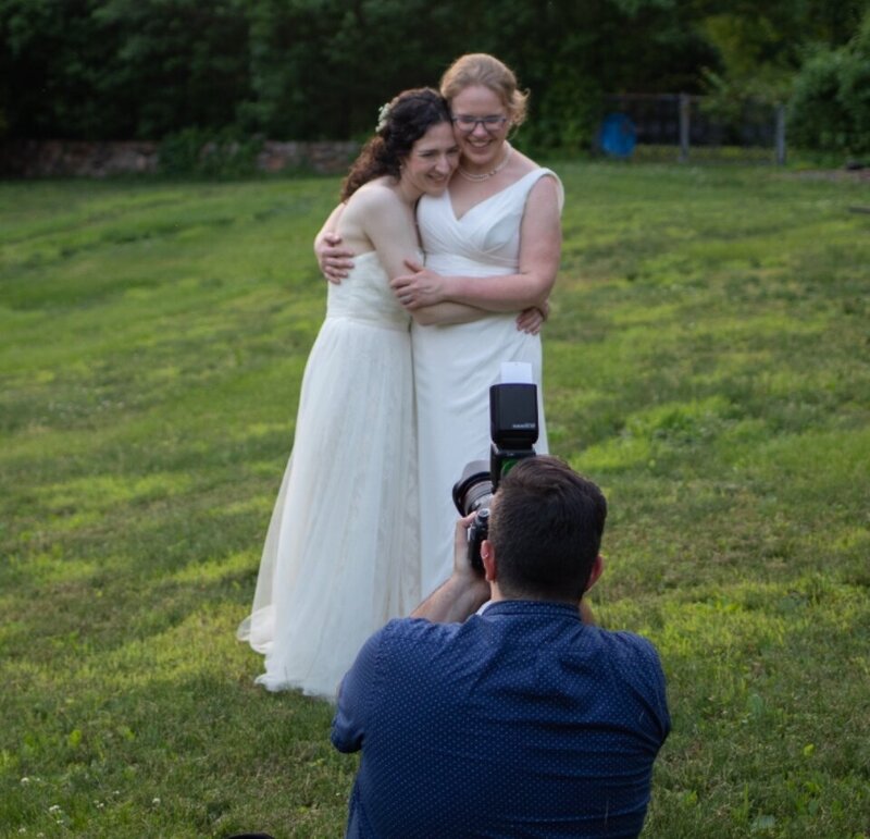 A photographer kneeling down as he captures an image of two brides hugging.