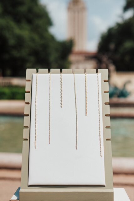 Selection of delicate gold-filled enamel chains in neutral tones displayed vertically on a jewelry board with a fountain in the background.