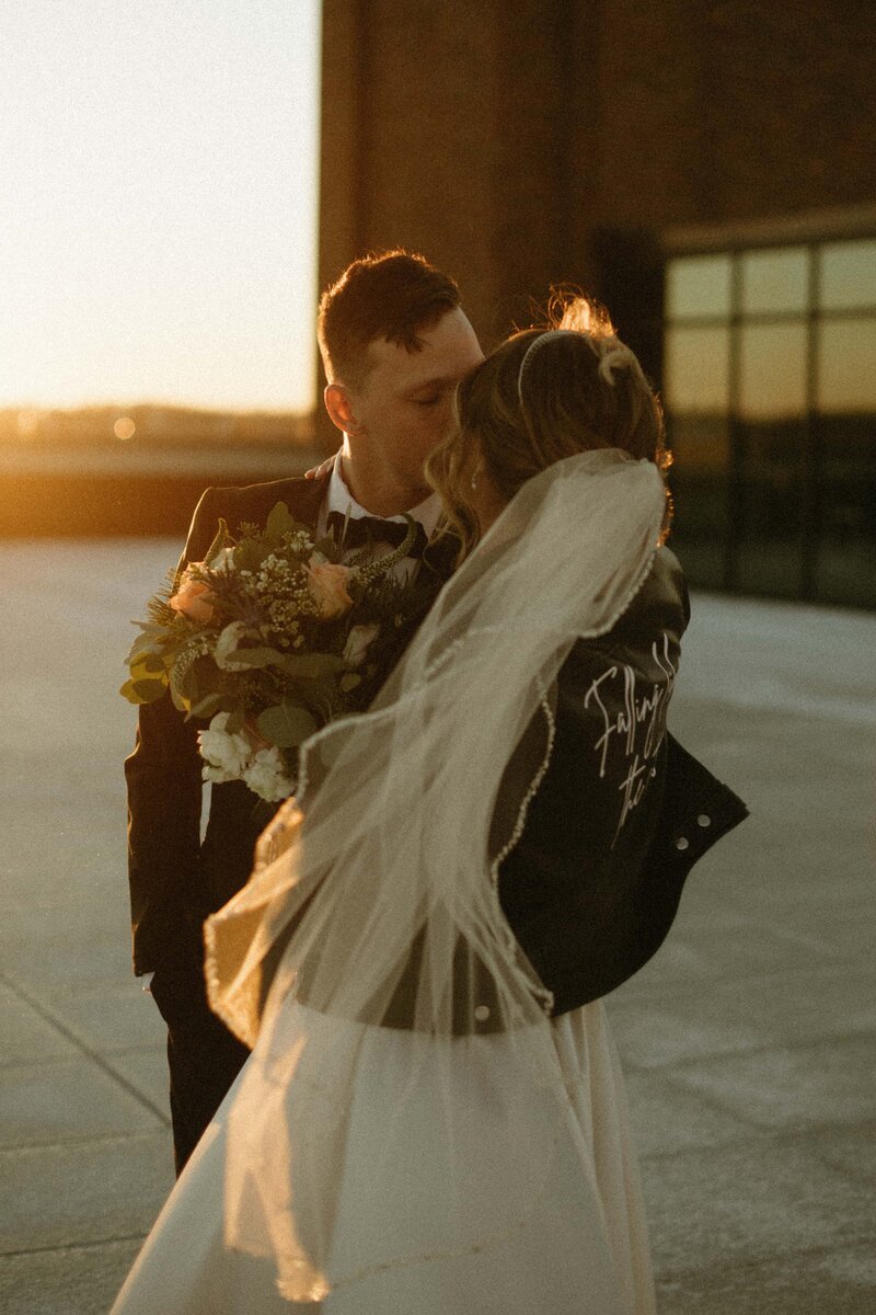 A bride and groom kissing at a park farm winery wedding, the bride in a white gown and veiled head holding a bouquet, the groom in a black suit, at sunset.