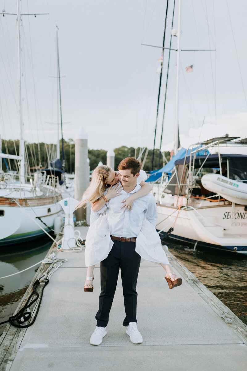 Bride in white cotton dress and brown wedges is riding on grooms back and peaking around so they look at each other walking down a dock with white sailboats on each side.