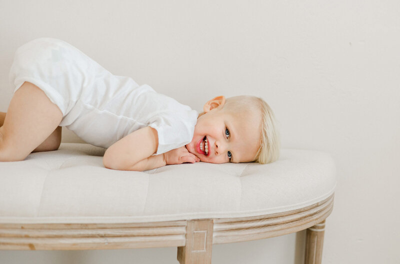 A child in a white onsie smiling a the camera while laying on a  linen bench in Daniele Rose Photography's studio