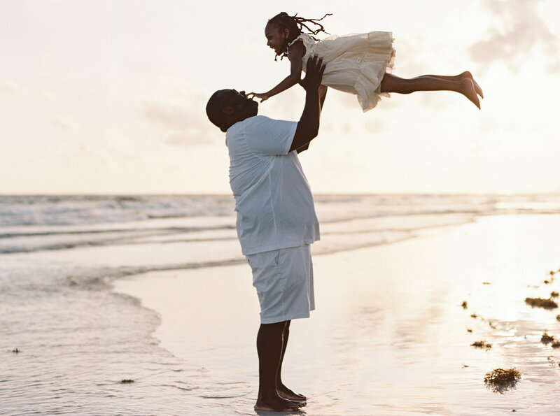 A father throwing his daughter up in the air at the beach while the sun sets a golden glow.