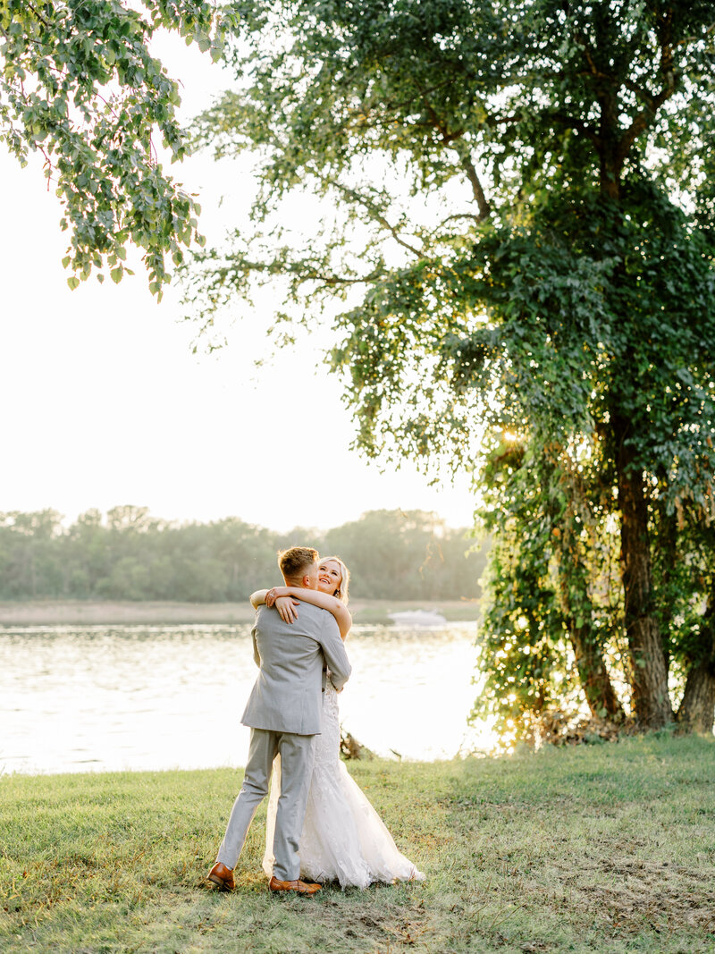 Bride and groom with their arms around each other with the sun setting on the water behind them