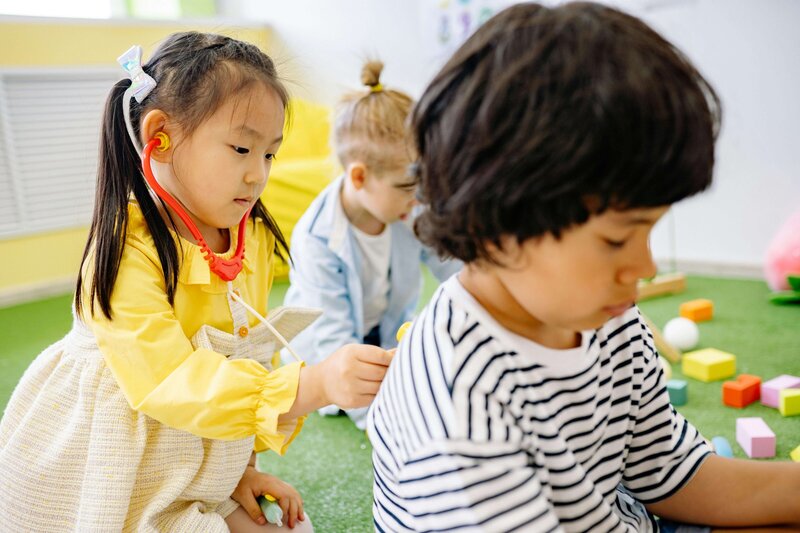 A young girl wearing a stethoscope pretends to examine a boy's back while another child watches in a playful, pretend-doctor activity.