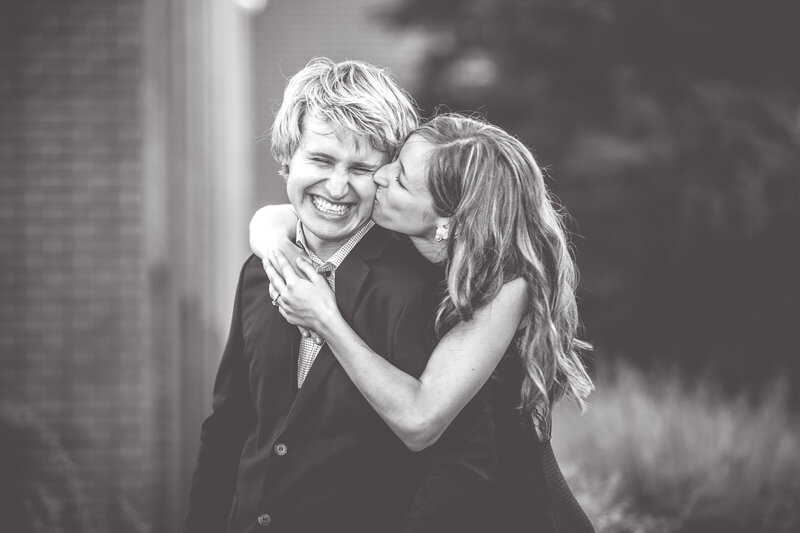 A cute photo of a bride kissing her groom while he is laughing during an outdoor engagement session.