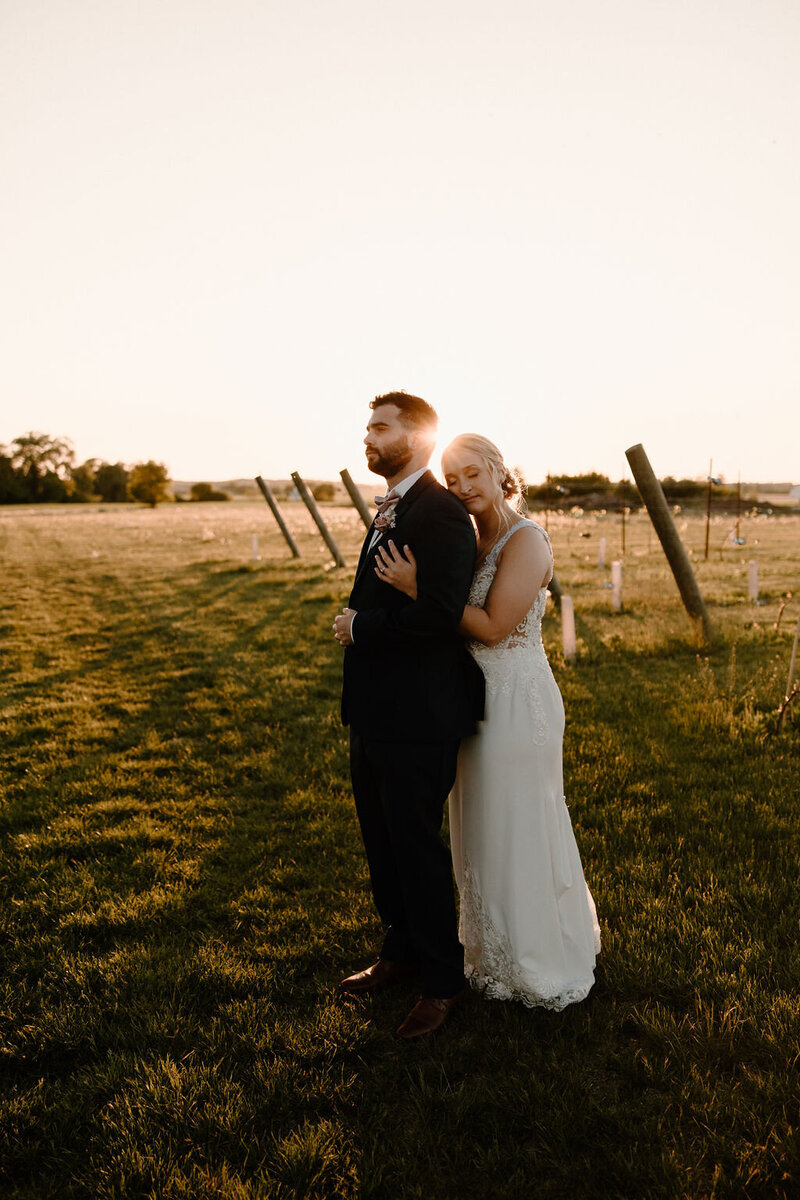 Beautiful, dark and moody wedding photography byMorgan Ashley Lynn Photography in Lake Country, Wisconsin out in a field at sunset with bride hugging the groom from behind and looking at the camera