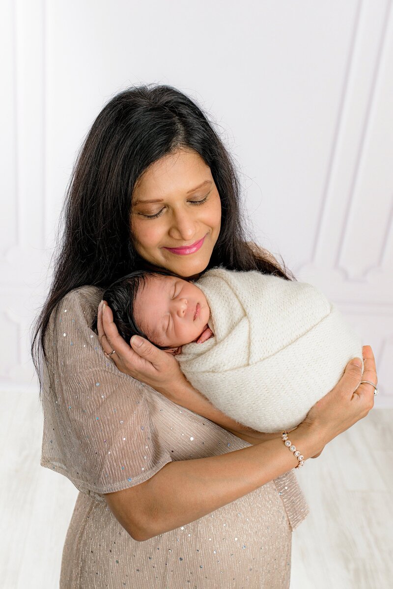 Mother is dressed in a champagne dress holding her newborn son wrapped in cream in a Charlotte Studio