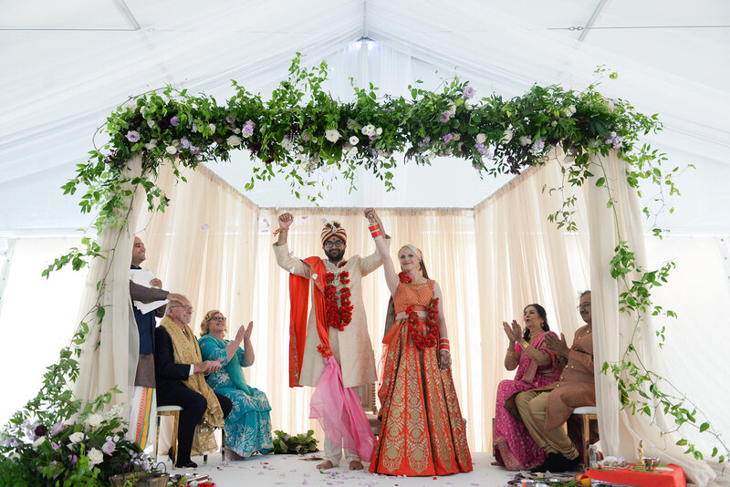 An Indian man, white woman and their immediate families celebrate the couple's wedding  ceremony under a stunning traditional mandap featuring taupe chiffon draping and a large garden-inspired mauve, lilac and plum coloured fresh floral installation along the top.