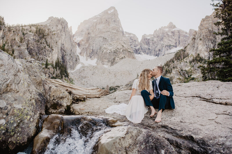 jackson hole photographers photographs delta lake elopement with bride and groom sitting on a rock together in the tetons as the groom kisses the woman head