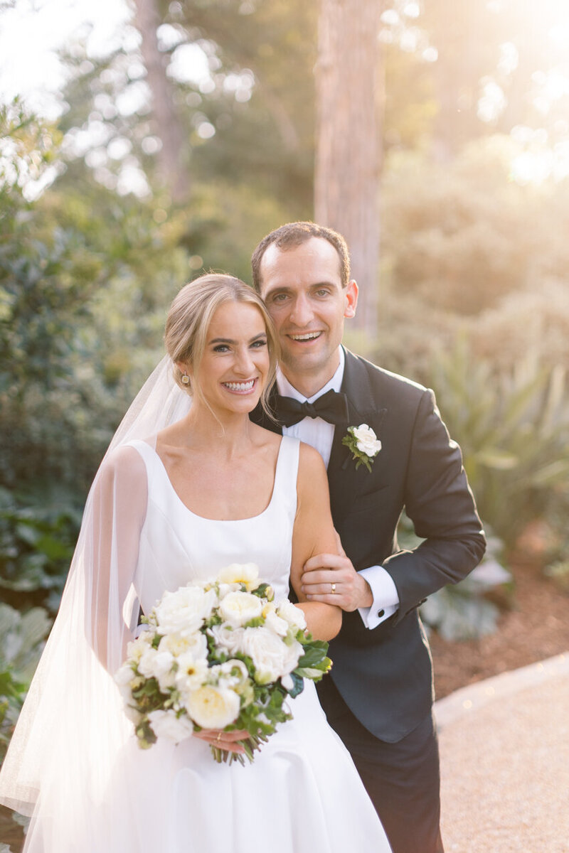 A bride and groom smile at the camera, there is a sun glare behind them. The bride's satin dress has a scoop neckline and thick shoulder straps