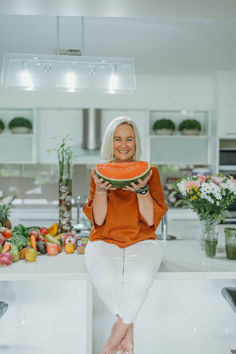 Creative branding photography of a woman in a bright kitchen holding a watermelon slice with fresh produce around her.