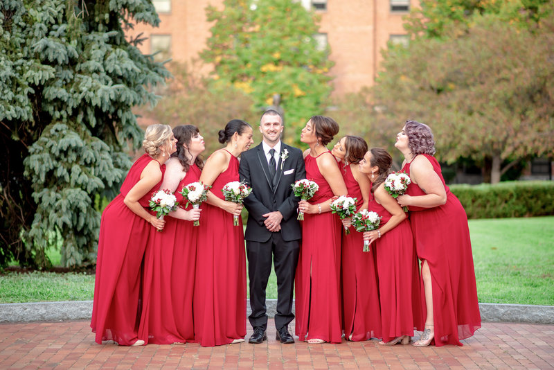 Bridesmaids in red dresses lean in for a kiss with the groom.