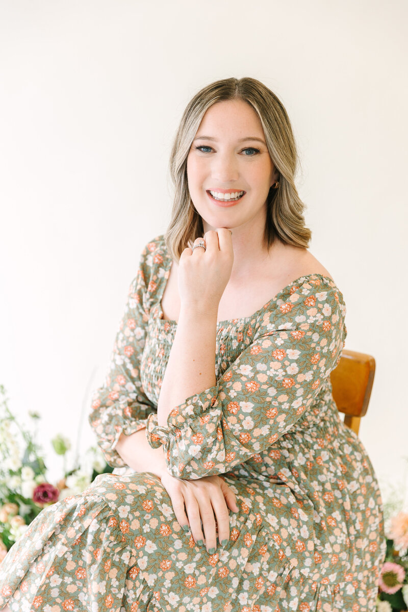 Amy Jordan of Amy Jordan Photography sitting in a studio wearing a floral dress and smiling.