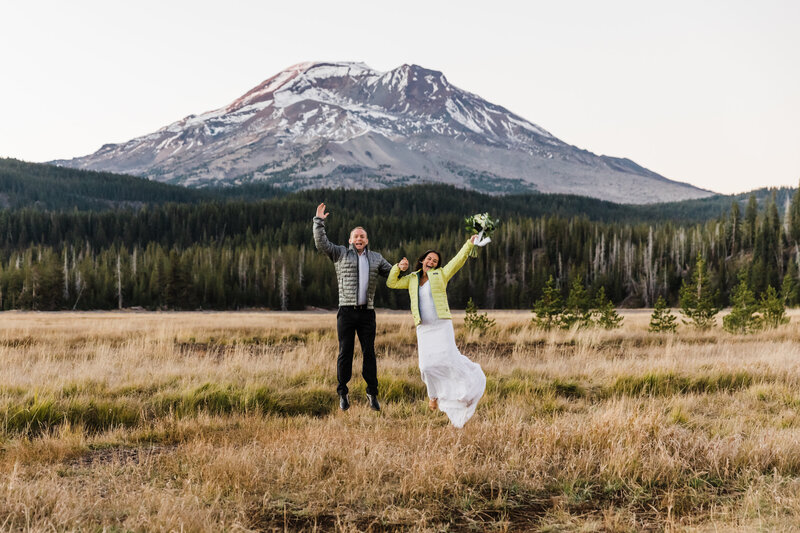 An Asian bride wearing a white dress and yellow Patagonia puffy jacket stretches her bouquet in the air with one arm while holding the hand of her groom wearing a white button down shirt and black slacks with a grey Patagonia puffy jumping in a meadow of dried grasses in front of South Sister at their adventure elopement near Bend, Oregon. | Erica Swantek Photography