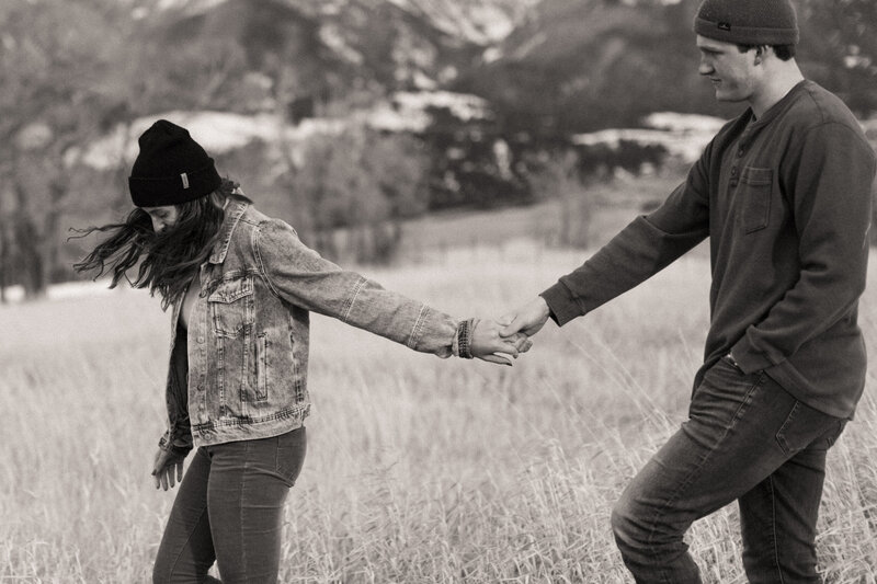 Couple wearing beanies holding hands walking through field in valley with snowy mountains in background