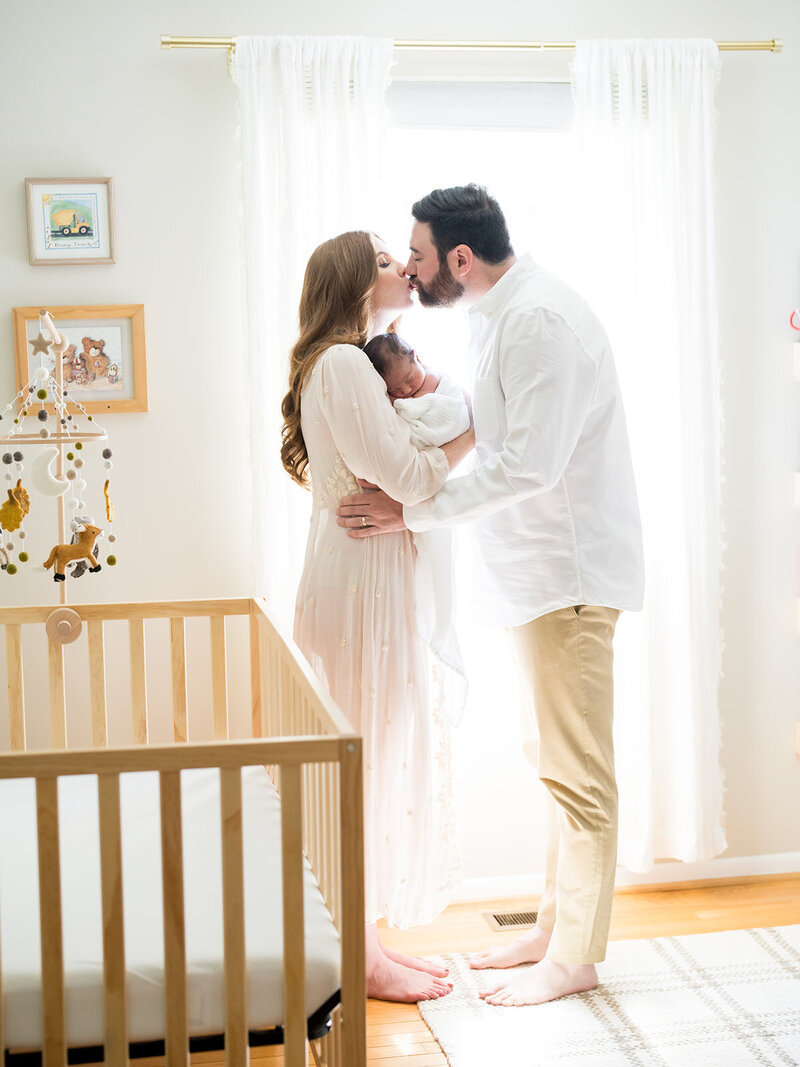 Parents lean in to kiss as they stand in front of the nursery window holding newborn baby during their Annapolis newborn photography session.