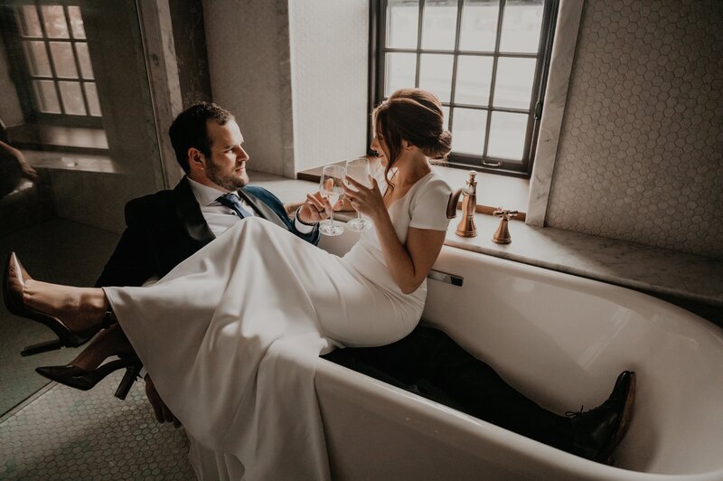 Bride and Groom pose for intimate wedding portraits. Groom is wearing is suit sitting in a claw-foot tub with his bride in her gown sitting on his lap with her legs over the edge of the tub. Bride and Groom are clinking champagne classes and smiling at each other. Image captured by Ashlee Ellison Photography, top Toronto, ON wedding photographer and videographer