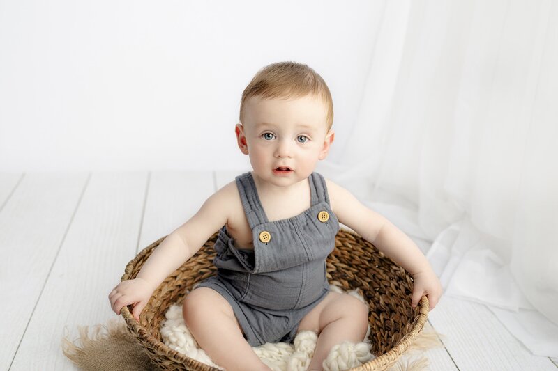 little boy holds onto the edge of a brown basket as he sits in the middle