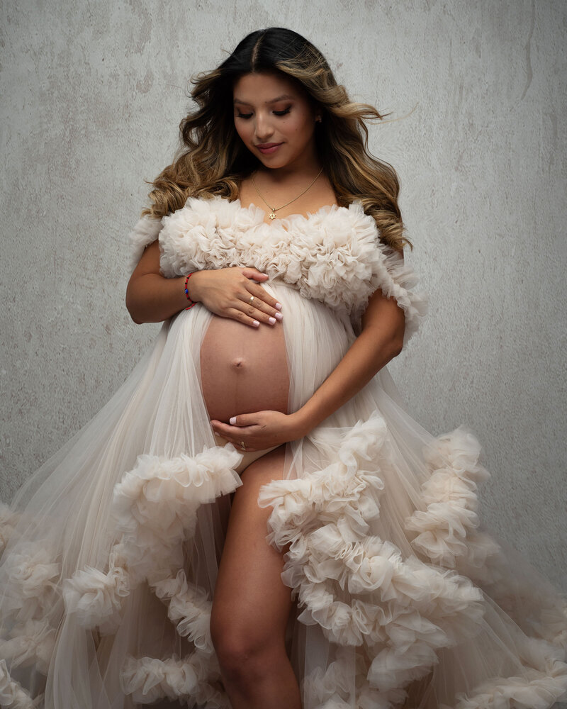 Pregnant woman looking down holding belly with tan skin wearing cream tulle coco gown by Kathatrina Hakaj with belly bump  and leg showing