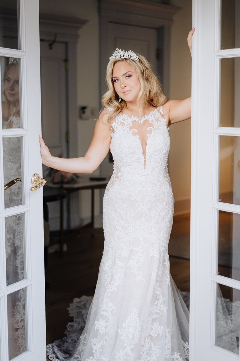 A bride in a lace gown and tiara stands confidently in a doorway, reflecting the grace and sophistication of Socially Significant Events’ wedding planning
