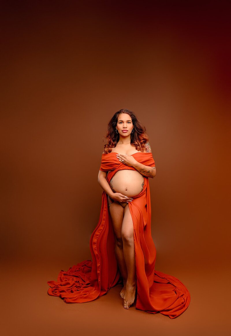 pregnant mother is wrapped in a rust chiffon with belly and leg exposed and both hands on her belly on a charlotte studio background